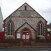 Front view of a tiny, plain chapel of irregular flintwork and red brick dressings. A dark stone plaque below a slit window near the roofline reads "WESLEYAN CHAPEL".  An entrance porch is flanked by two wide, low-set windows with pointed arches.