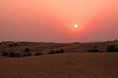 View of dunes at sunset. The setting sun hangs low and dim in a sky graded from fiery orange at top to maroon near the horizon. Terrestrial features are difficult to discern in the crepuscular illumination.