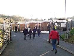 Various people walking towards the entrance to a brick-built sports stadium.