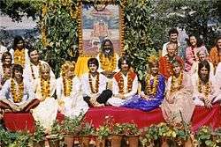 Formal portrait, taken outdoors, with people sitting cross-legged on a red platform in front of a large painting.