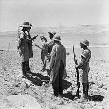 Two British soldiers face right while they talk to two French soldiers (centre). A third British soldier stands in the background, and a fourth stands on guard behind (to the left) of the French troops.