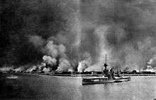 A city burns in flames; in the foreground, a large battleship sits in the water. A smaller vessel is berthed next to her. Another small ship sails away from the city on the left.