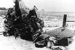 A tangled mass of metal with a wing and landing gear wheel barely recognizable, on a snowy field