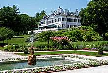 A white mansion house stands in a garden. A terraced lawn separates the house from a fountain with a pool fringed by white plants.