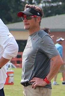 Candid waist-up photograph of Dimitroff wearing a grey t-shirt and black visor both bearing Atlanta Falcons logos and standing on a football practice field with arms akimbo