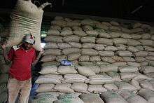 A man carries a large bag on his shoulders.  Behind him, thousands of bags are stacked in rows.