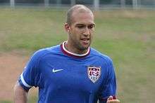 A photograph of a bald mixed-race man wearing a royal blue shirt.