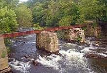 A rusted steel girder supported by two stone piers in the middle of a fast-flowing stream in a wooded area