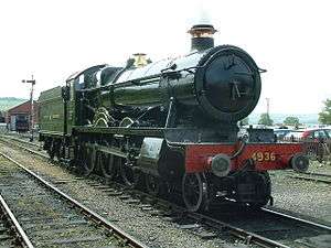 A locomotive is seen from in front and from the right. The locomotive is mainly green but the smoke box (at the front) is black and the buffer beam is red with the number 4936 written in yellow letters. The chimney is trimmed with a copper ring and the valve cover is made of brass. Behind the locomotive is a green tender with the words "GREAT WESTERN" written in yellow. In the background to the left is a signal and an engine shed with a tank engine, and to the right is a car park behind a wooden rail.