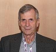 An older man in a dress suit standing in front of a wood background.