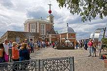 A group of people waiting in a line curving to the left on a cobblestone surface. Behind it is an ornate brick building with a red ball on top. The people at the end of the line, closest to the camera, are taking pictures of other people near a shiny metal monument on the right, under a tree. A line in the cobblestone connects them