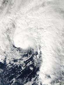 Satellite image of a comma-shaped tropical cyclone just off the United States Gulf Coast. All land in the image is obscured by cloud cover which extends in all directions, except southwest, from the center of the storm.