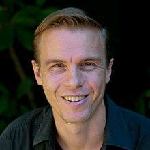 A portrait photograph of Troy Hunt's head and shoulders. Hunt has light skin and brown hair, which is short and slicked back. He is looking directly at the viewer and smiling with his top row of teeth showing. He is wearing a dark blue shirt, and is against a dark green and black background.