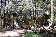  A large polygonal wooden building sits in a sunny second-growth forest. A woman is kneeling in front of a wooden bench along a path. A baby stroller is parked next to the bench. A few feet away from her, three small children and another woman stand on the path.