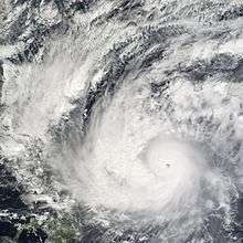 A well organized tropical cyclone, centered at bottom right, with a well-defined eye and spiraling rainbands. A landmass is visible under heavy cloud cover at left.