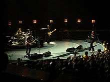 A colorful, round stage inside an arena, with all four of the band members performing their instruments.