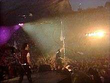 A shot of a concert stage from the back at dusk. On the left, The Edge plays guitar. In the center, Bono, at the edge of the stage, holds up a white flag near audience members, several of whom help him support the flag. In the center of the audience are bright lights aimed in the direction of the performers. Behind the audience are large red cliffs. Red streaks of light are visible throughout the image