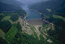 Aerial view of a dark blue lake, surrounded by forest, with a dam in front of the lake