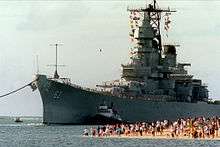 A large gray ship moves toward the camera and slightly to the left. On the right, a number of people on a beach are observing the ship as she moves in to pass them with the aid of a tugboat.