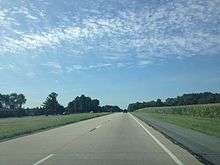 A four lane concrete road with a grassy median surrounded by farms and woods