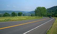 A paved two-lane road crosses the lower half of the picture from left to right. A small blue car is visible at the right with its brake lamps on, where the road turns back sharply. After the turn it is slightly downhill from the camera and cannot be seen, although black-on-yellow arrow signs indicate where it is. In the distance, in the top of the image, is a valley with green hills on either side