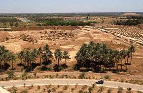 From the foot of Saddam Hussein's summer palace a Humvee is seen driving down a road towards the left. Palm trees grow near the road and the ruins of Babylon can be seen in the background.