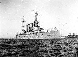 An armored cruiser anchored, with a navy jack flying from the jackstaff.