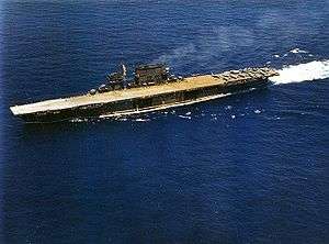 left front oblique aerial view of a dark-gray aircraft carrier at sea. Her flight deck is beige colored and has a dozen or so aircraft at the stern.