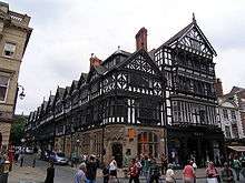 The corner of a street with a range of shops running up the right side of the street; the shops have stone lower storeys and highly detailed timber-framed upper storeys that include oriel windows and gables. To the right of the range of shops is another shop with a modern shop front; it is taller than the others, and is timber-framed above the ground floor.