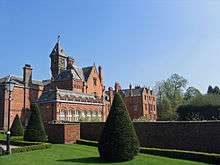 An irregularly shaped red-brick house with slate roofs with an orangery on the ground floor and gables, chimneys and a clock tower above.  In the foreground is a formal garden with a lawn, clipped shrubs, a wall and a Victorian-style lamppost.