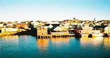 A cluster of buildings and wharves, with a small ship moored to the right of picture. The waterside buildings are clearly reflected in the sea.