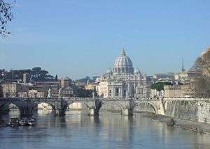 A view of Rome on a sunny afternoon looking along the river. A bridge crosses the river and beyond it is a hill on which the grey dome of St. Peter's rises above ancient buildings and dark pine trees.