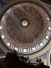  Photo looking up at the dome's interior from below. The dome is decorated at the top with a band of script. Around its base are windows through which the light streams. The decoration is divided by many vertical ribs which are ornamented with golden stars.