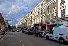 A street seen at angle with an attached row of four-story beige brick buildings running deep into the background on the right with cars parked in front of the stores, including Burger King and McDonald's, at street level. On the left a much smaller portion can be seen, up to a tower a block away