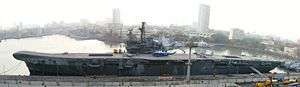 A small aircraft carrier tied up at a wharf. A helicopter and two aircraft sit on the flight deck. Other ships and skyscrapers are in the background.