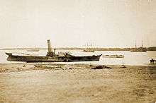 Photograph showing the hulk of a side-wheel steamship sitting very high in shallow water and heavily listing adjacent to a beach with other ships in the background