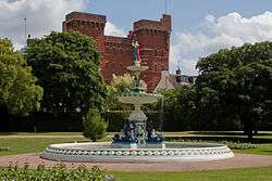 Ornate circular fountain surrounded by grassy area with trees and red brick building in the background.