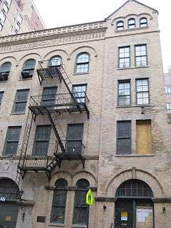 The right half of an old, sturdy-looking 4-story, 5-bay building, faced with partly soiled beige brick and architectural decorations. The right bay has a gabled fifth floor with 3 small windows.  A fire escape runs down the front. At street level 1½ large doorways are visible. One of the 20 or so windows is boarded, another is painted black.