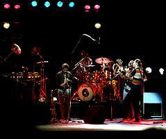 Four men performing various instruments on a stage, surrounded by a drum set and other audio equipment