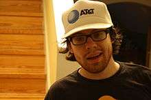 Portrait of a young man with long dark hair, stubble, spectacles and a white baseball cap