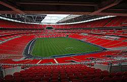 The interior of an empty stadium as viewed from its upper tier of seating. The seats are a vivid red and the pitch is a vivid green. The pale grey sky is visible through an opening in the ceiling above the pitch.