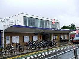 A brown-bricked building with a rectangular, dark blue sign reading "WEST RUISLIP STATION" in white letters all under a light blue sky