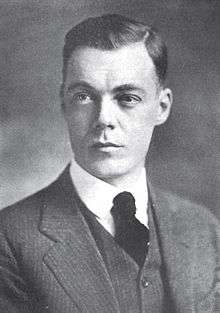 A young, dark-haired man wearing a black jacket, vest, and tie and a white shirt