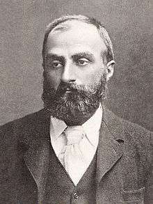  Heavily bearded man with dark receding hair, wearing a dark coloured jacket, white collar and pale tie. He is looking slightly to the left, with a solemn expression