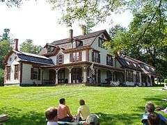 A large ornately-decorated country house. It is painted a light color with brown trim, and the porch has detailed decoration painted the body color. There are small round windows above some of normal windows. People sit at tables on the porch that wraps part way around the house, and also on the expanse of lawn in front.