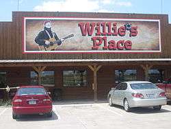 A store with a sign that reads "Willie's Place". The apostrophe is replaced in the sign by a bullet hole. The structure of the store is constructed in wooden with three columns. There are four windows and there are a red and a grey car in the parking lot.