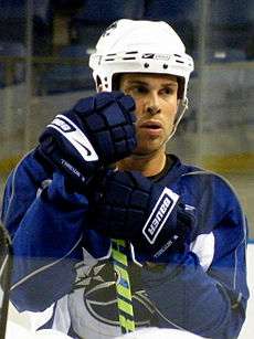 A Cacucasian ice hockey player leaning on his hockey stick. He wears a blue helmet and jersey.