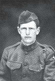 Head and shoulders of a stern faced man in a military uniform with breast pockets, shoulder straps, and garrison cap.