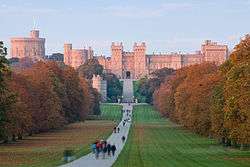 A red castle with battlements and towers lies in the distance of the photograph. A path curves from the bottom of the picture towards it, with various people strolling along it. On either side is flat grass and green woodlands.