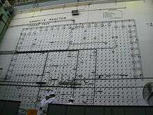 Two workmen on a movable platform similar to that used by window washers, in front of a wall with arrays of holes and many wires running across it. A sign says "Graphite Reactor loading face".